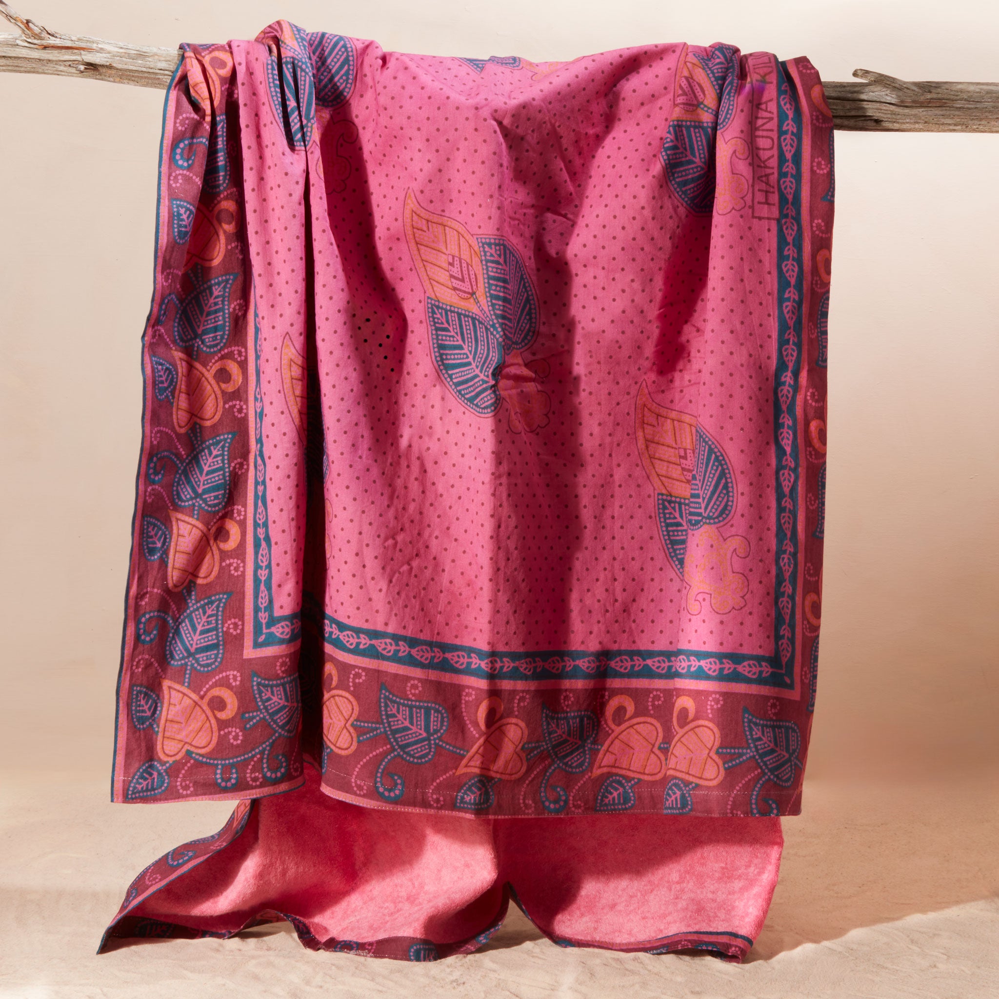 A pink kanga towel with one fabric side featuring a fine dotted pattern, a blue border, and orange and blue leaf motifs with a pink terry cloth towel lining.