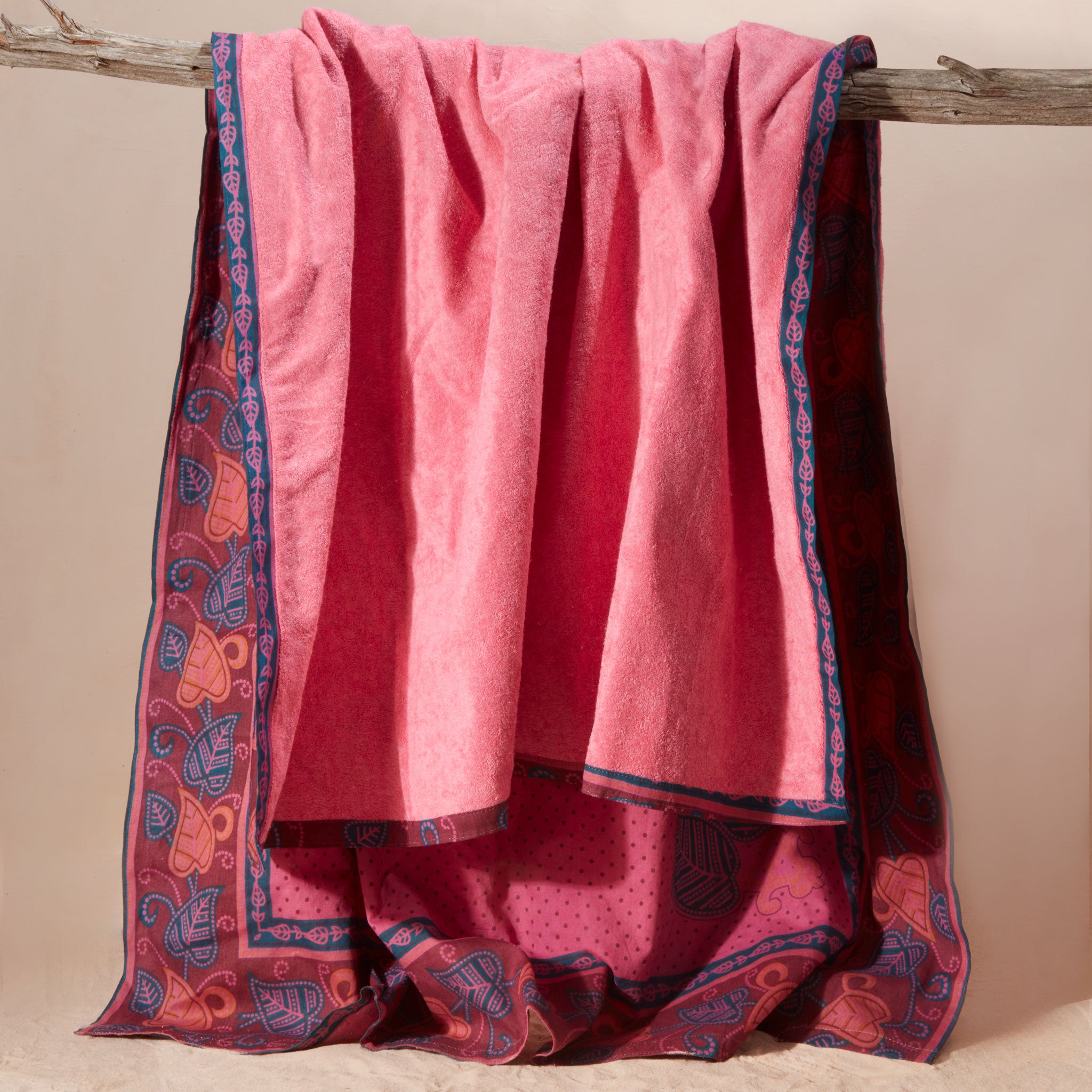 A pink kanga towel with one fabric side with a fine dotted pattern, a blue border, and orange and blue leaf motifs featuring the pink terry cloth towel lining on a rustic branch against a beige background.