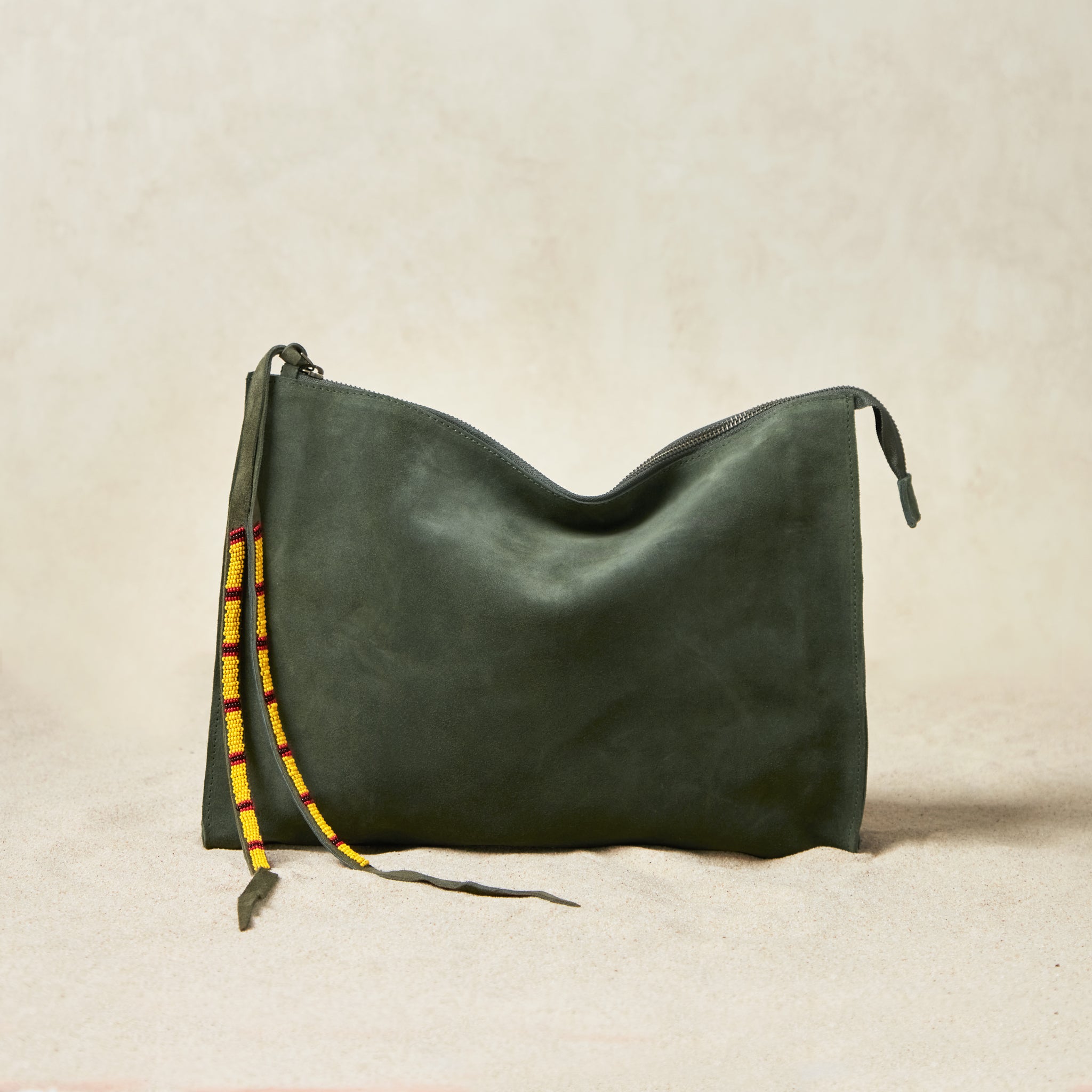 Large olive green suede leather pouch bag with yellow, black, and red beaded zipper pull ties on a sand background