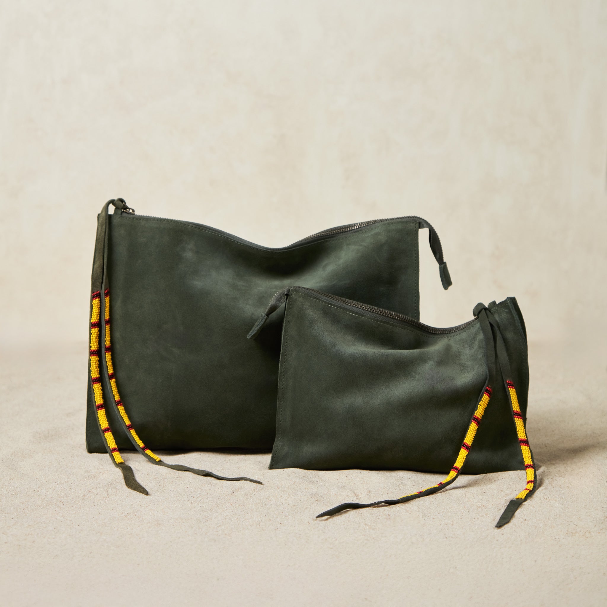 Two olive green suede leather bags in different sizes with yellow, black, and red beaded zipper pull ties on a sand background