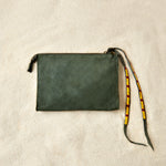 Flat view of small olive green suede leather pouch bag with yellow, black, and red beaded zipper pull ties on a sand background