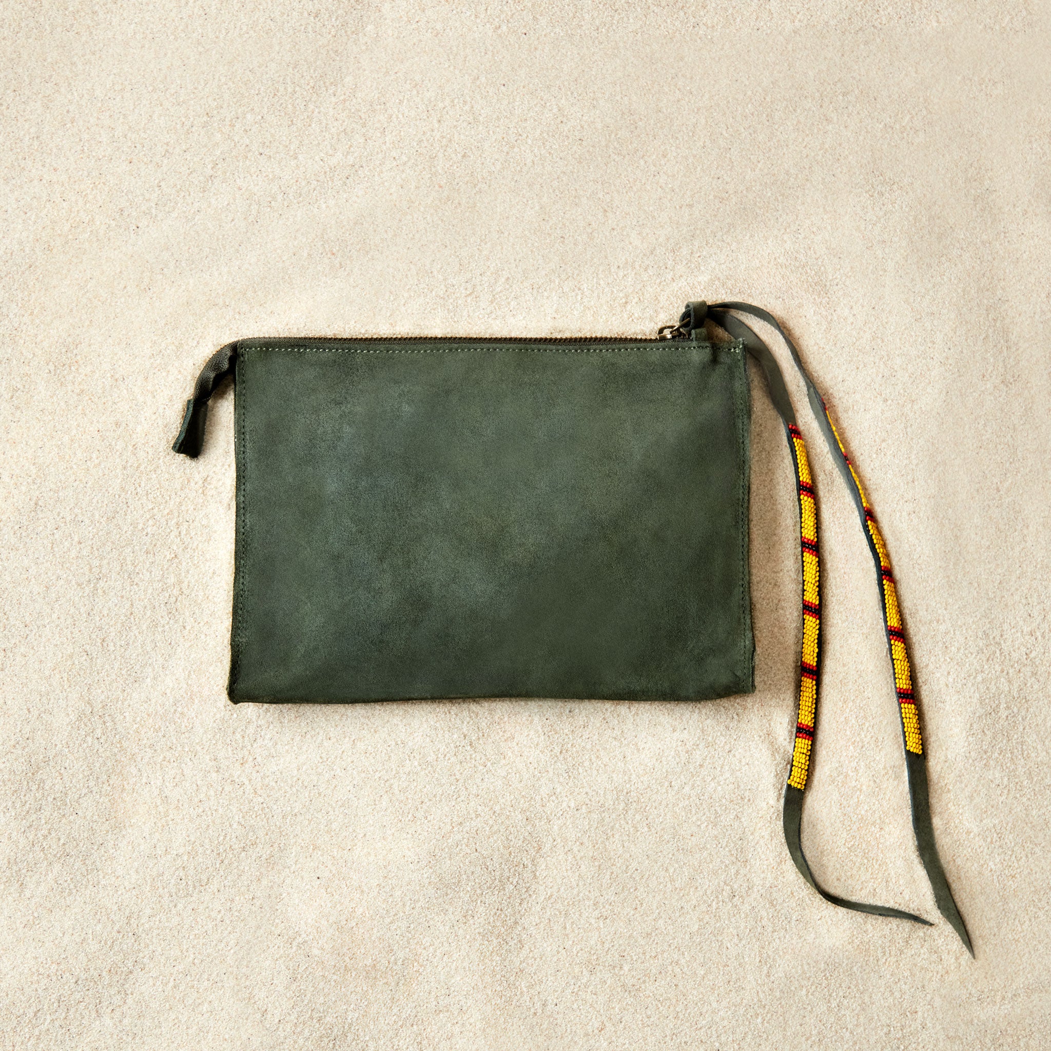 Flat view of small olive green suede leather pouch bag with yellow, black, and red beaded zipper pull ties on a sand background