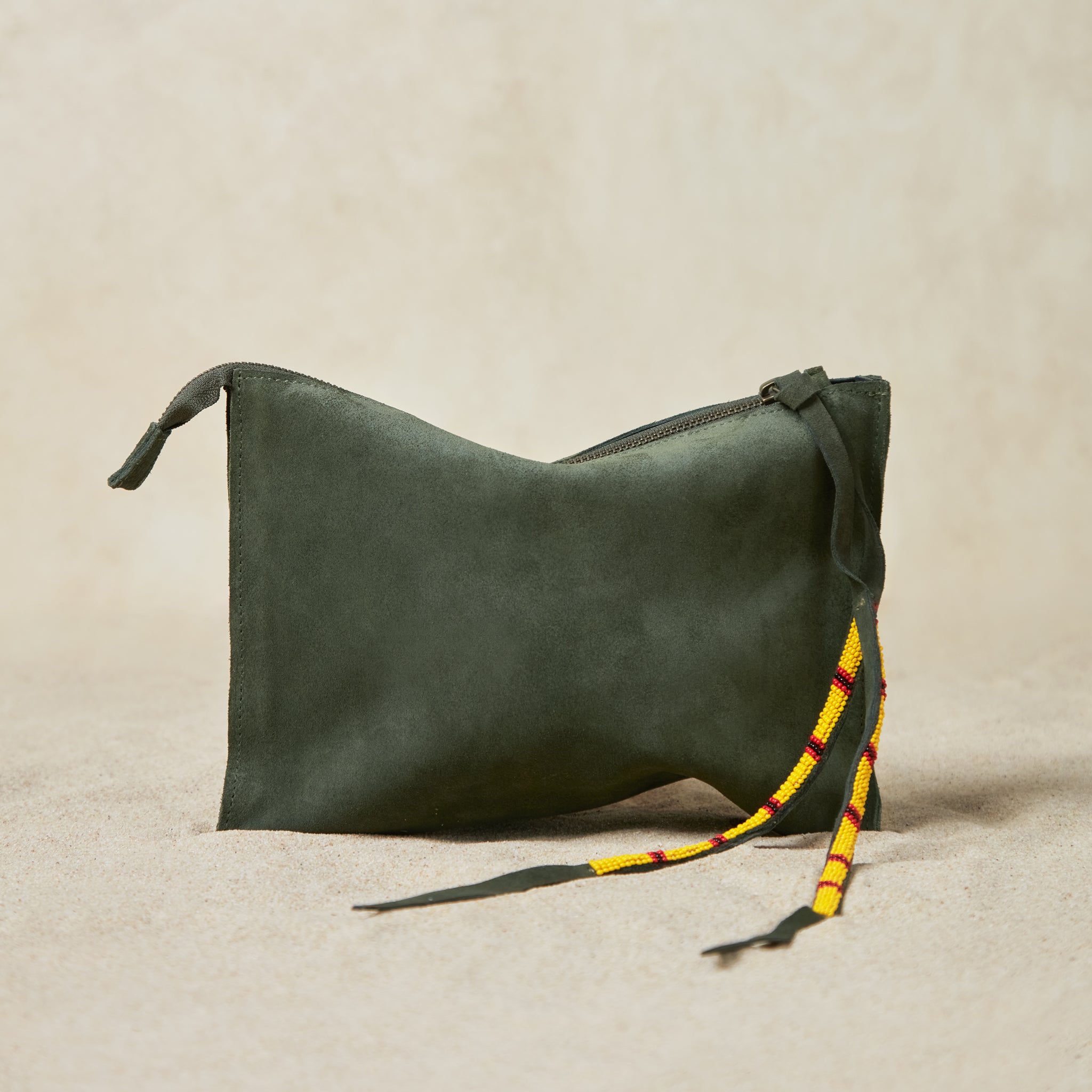 Small olive green suede leather pouch bag with yellow, black, and red beaded zipper pull ties on a sand background