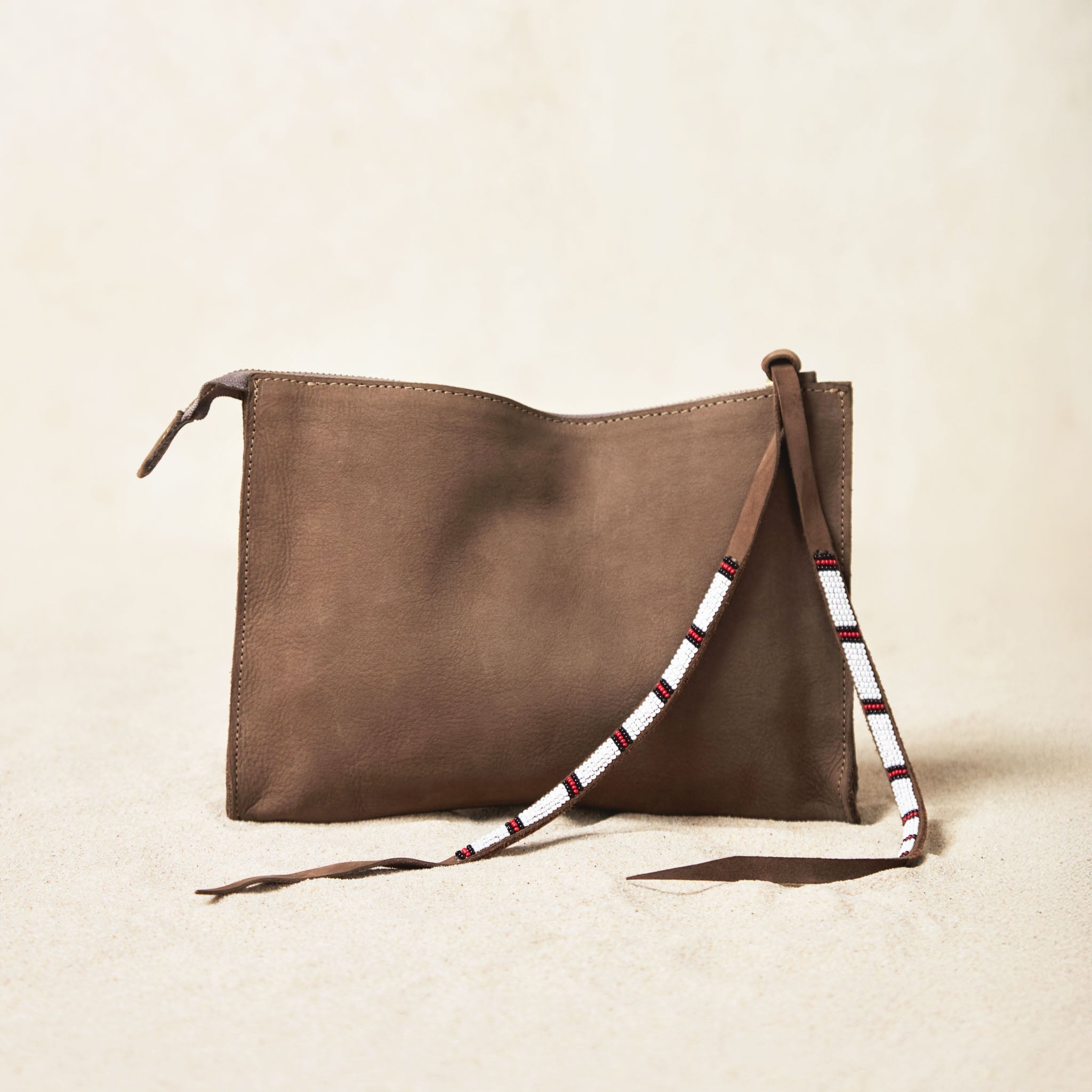 Small sand brown suede leather pouch bag with white, black, and red beaded zipper pull ties on a sand background