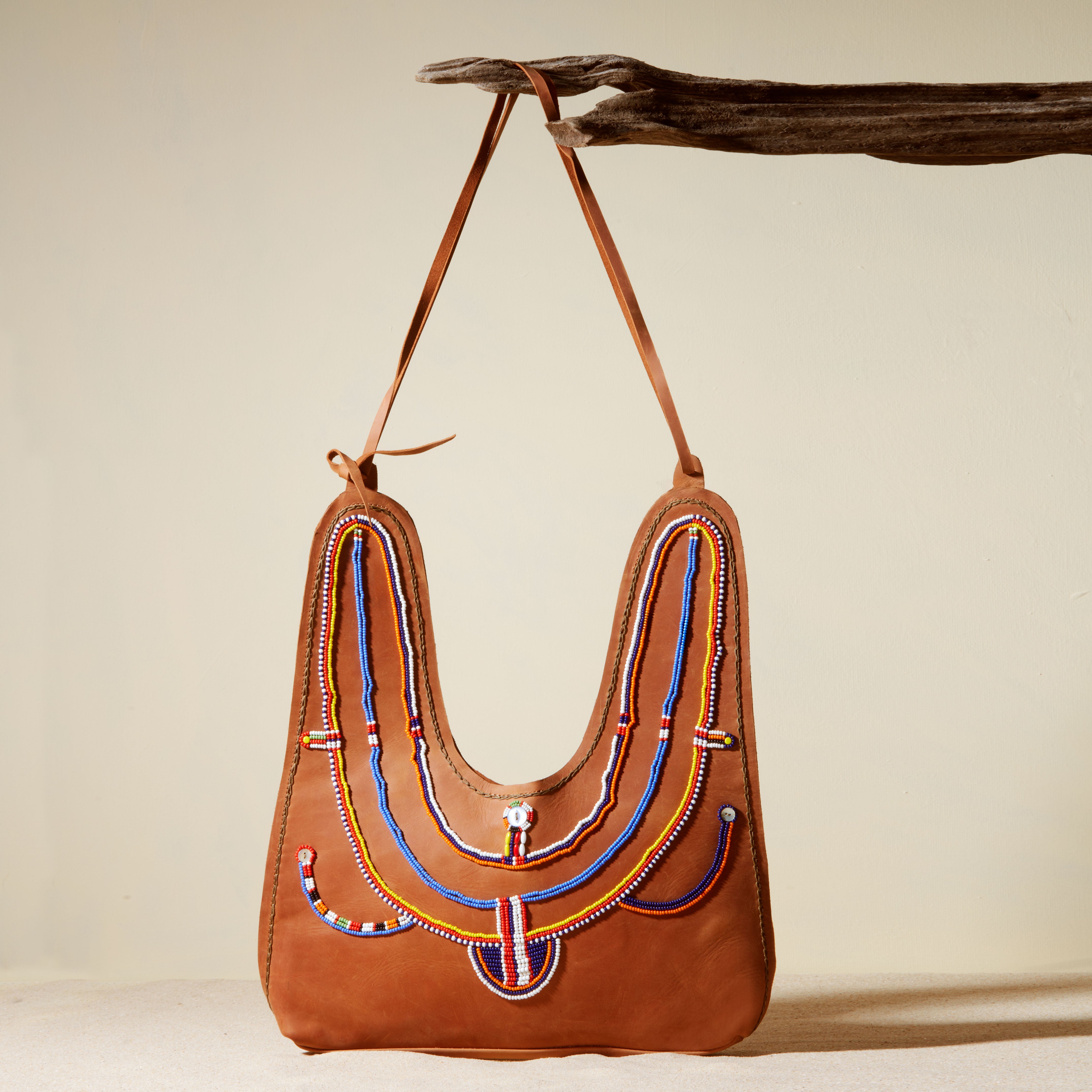 Brown leather handbag with colorful beadwork hanging on a branch against a beige background