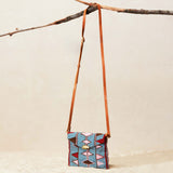 Small beaded crossbody bag with a colorful pattern and brown leather strap hanging on a branch on a beige background