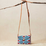 Small beaded crossbody bag with a geometric pattern and brown leather strap hanging on a branch against a beige background