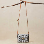 Small crossbody beaded bag with geometric pattern and a brown leather strap hanging on a branch on a beige background