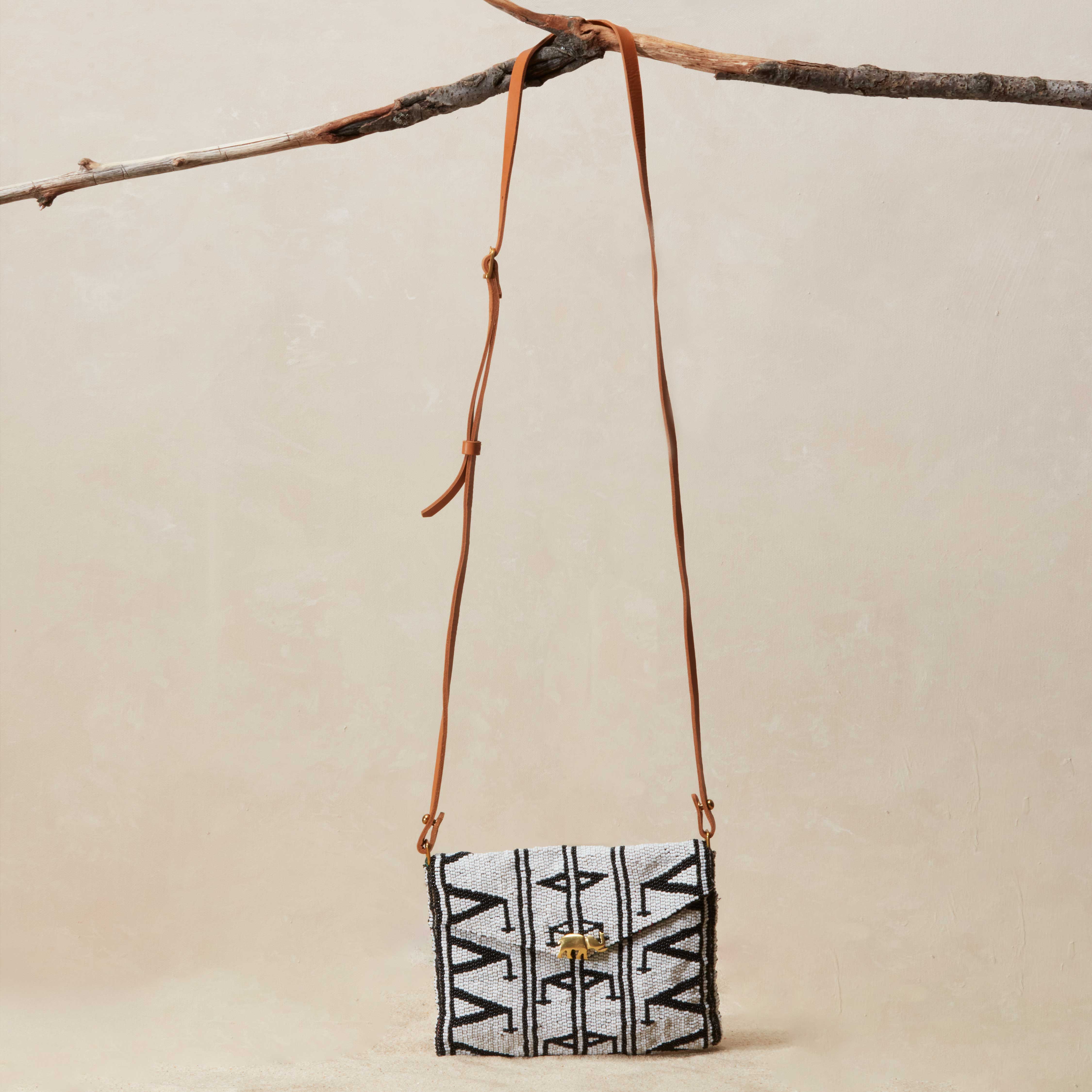 Small crossbody beaded bag with geometric pattern and a brown leather strap hanging on a branch on a beige background