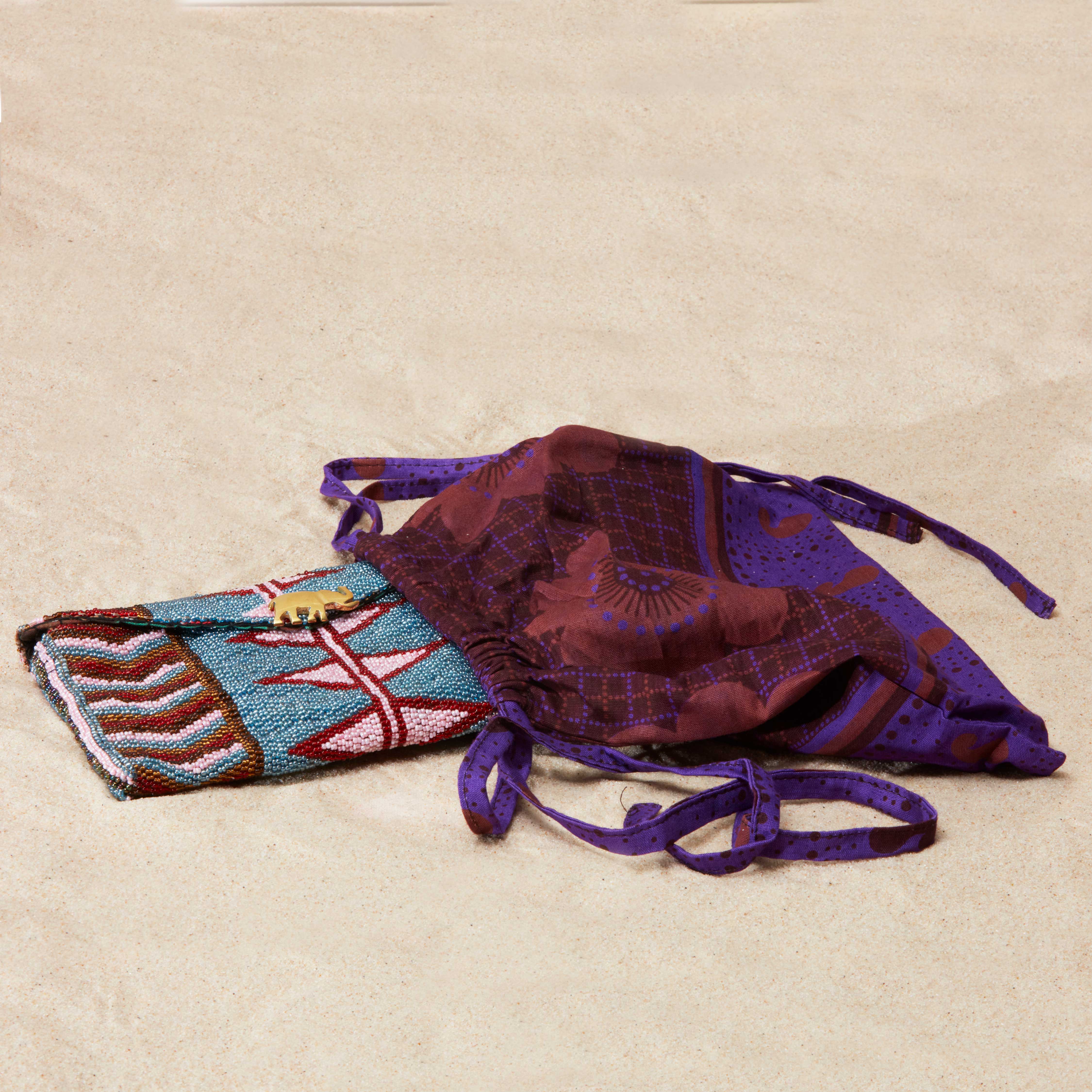 Colorful beaded clutch with brass elephant emblem inside a colorful purple kanga fabric dust bag on a sandy surface
