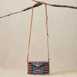 Beaded handbag with a geometric pattern and a brown leather strap hanging on a branch against a beige sandy background