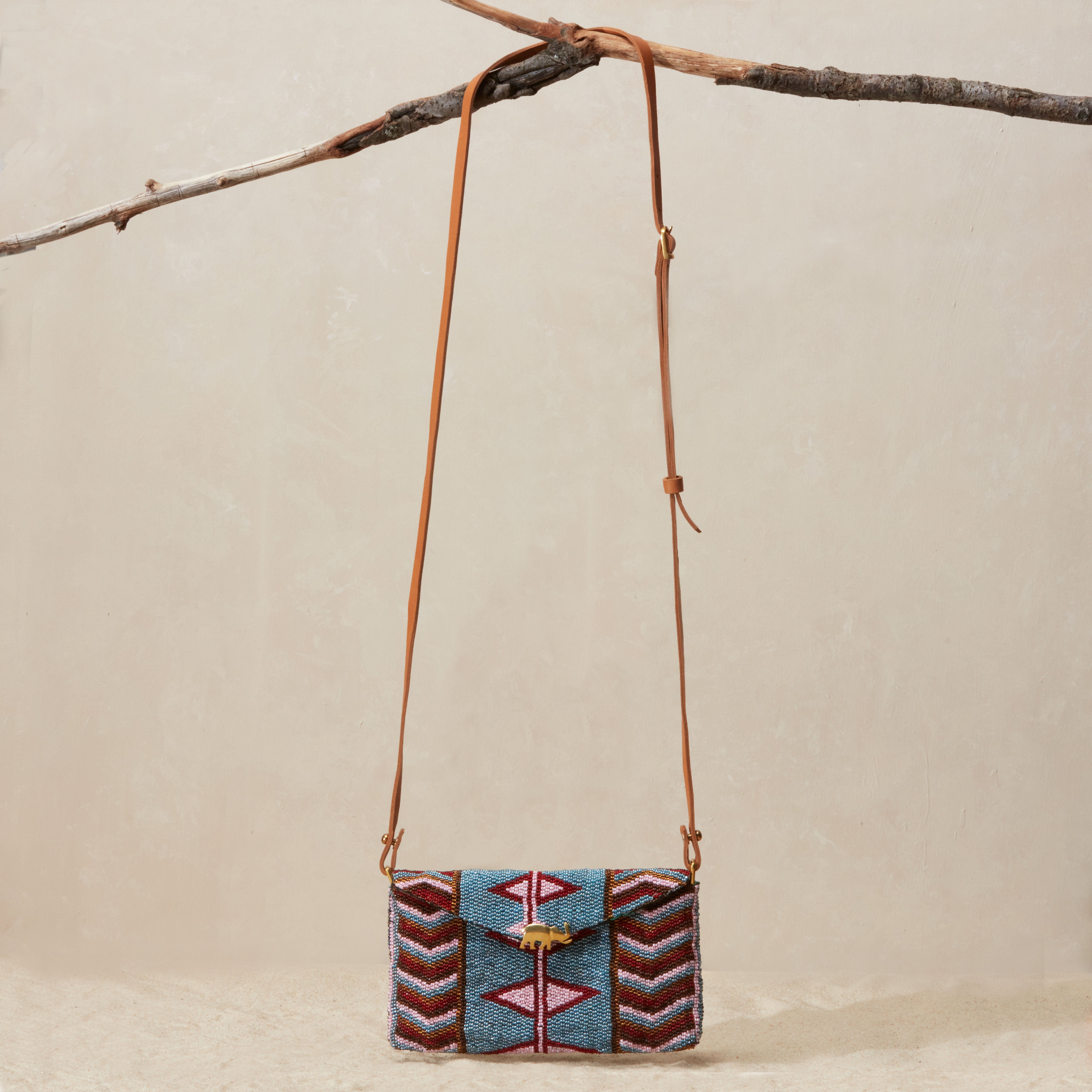 Beaded handbag with a geometric pattern and a brown leather strap hanging on a branch against a beige sandy background