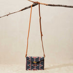 Small beaded handbag with geometric pattern and brown leather strap hanging on a branch against a beige background
