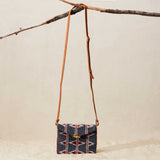Small beaded handbag with geometric pattern and brown leather strap hanging on a branch against a beige background