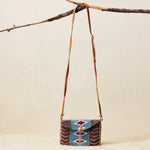 Small beaded handbag with a patterned design and brown leather strap hanging on a branch against a beige background
