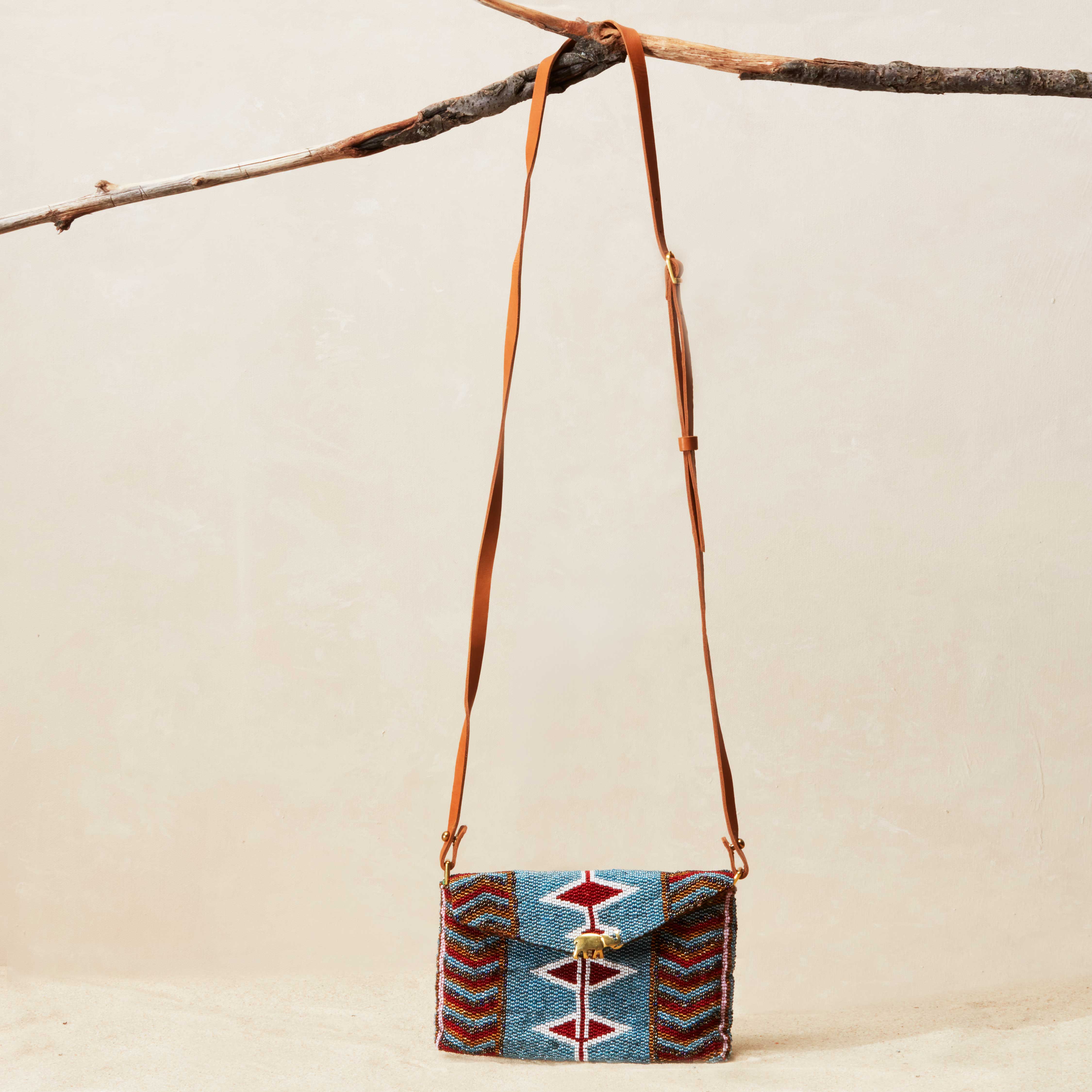 Small beaded handbag with a patterned design and brown leather strap hanging on a branch against a beige background