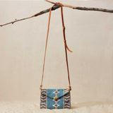 Blue beaded geometric handbag with brown leather strap hanging on a branch against a beige background