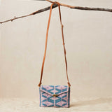 Beaded handbag with a geometric pattern and brown leather strap hanging on a branch against a beige background