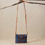 Blue and brown patterned beaded handbag with a brown leather strap hanging on a branch on a beige background