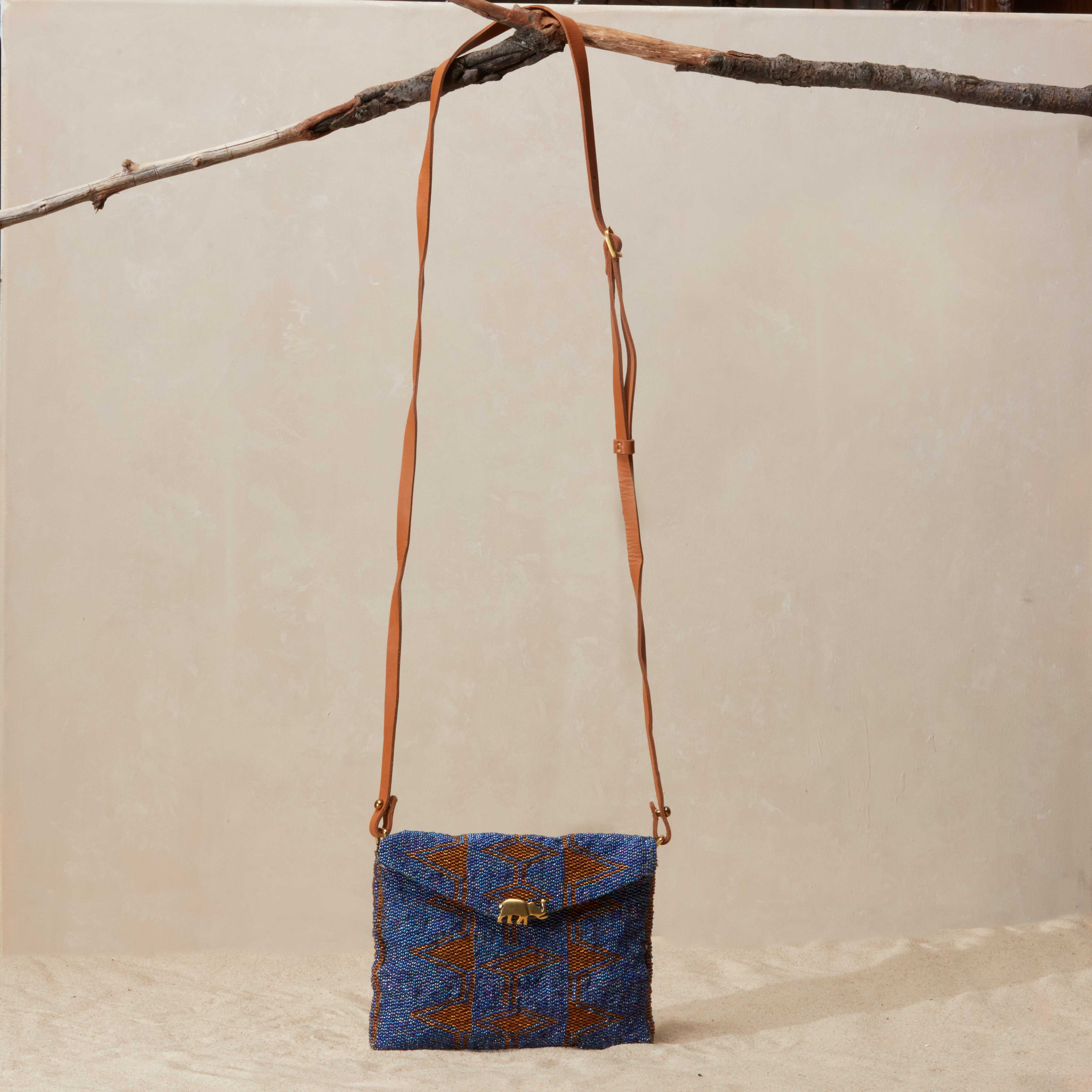 Blue and brown patterned beaded handbag with a brown leather strap hanging on a branch on a beige background
