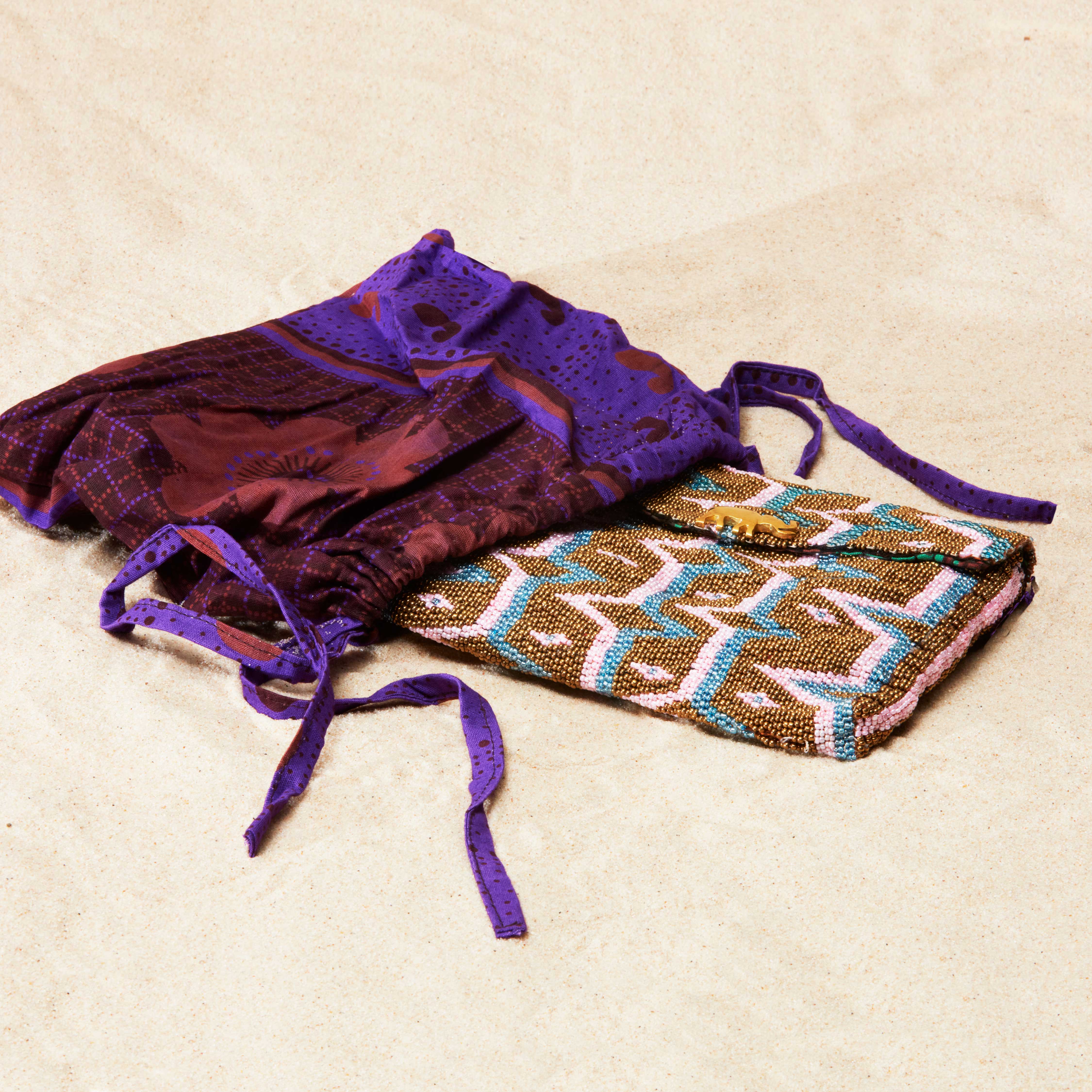 Colorful geometric patterned beaded clutch in a maroon and purple kanga fabric dust bag on a sandy surface