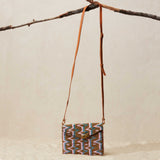 Small  beaded handbag with a patterned design and a brown leather strap hanging from a branch on a beige background
