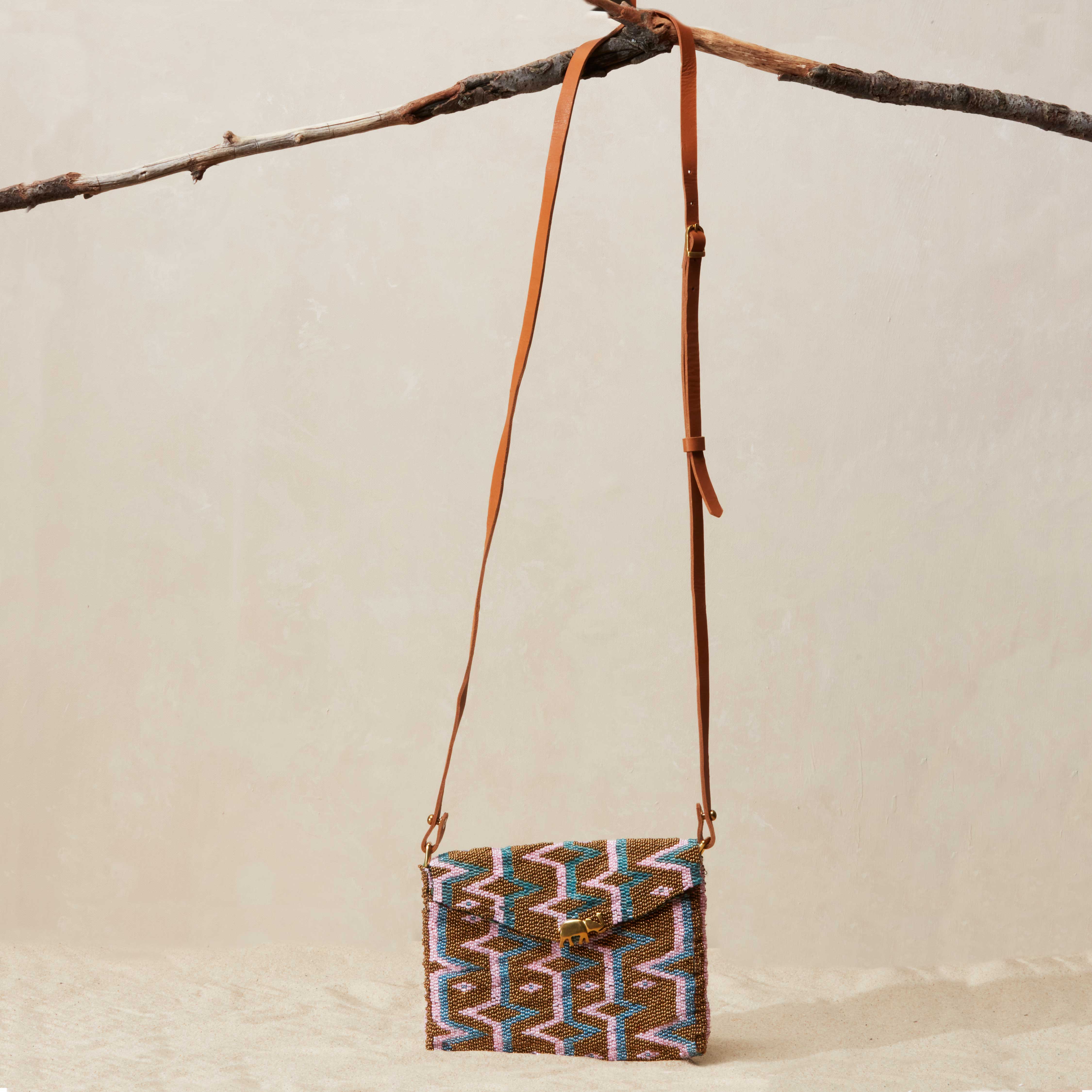 Small  beaded handbag with a patterned design and a brown leather strap hanging from a branch on a beige background
