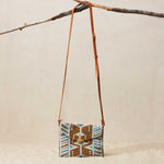 Small beaded crossbody bag with a  brown leather strap on a neutral background hanging from a branch