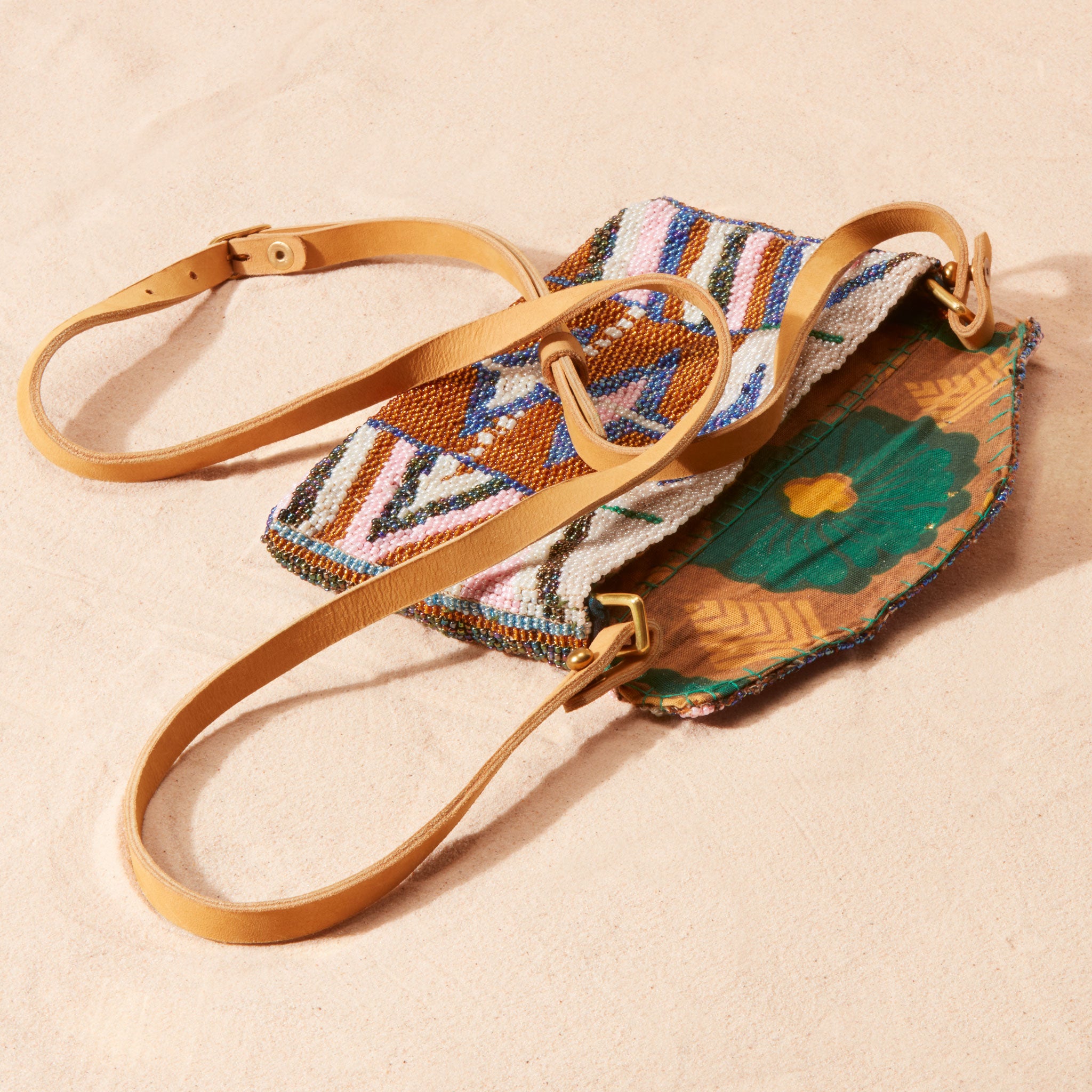 Colorful beaded clutch with brown leather strap, open showing the colorful kanga fabric lining of the bag on a sand background