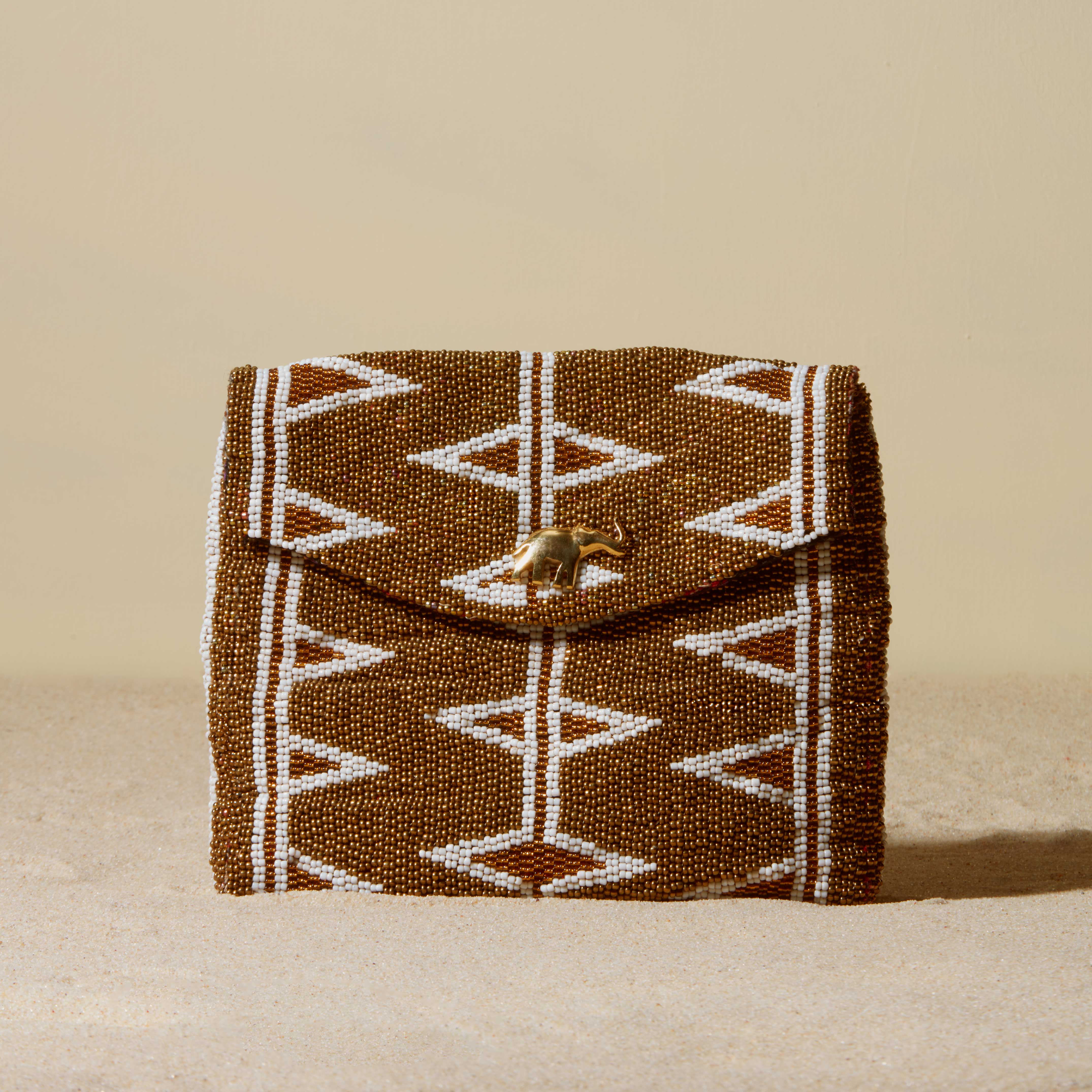 Gold and white beaded clutch with geoemtric pattern and a brass elephant on a sand background