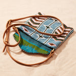 Beaded handbag with geometric pattern and brown leather strap, open showing colorful kanga fabric lining on a sand background