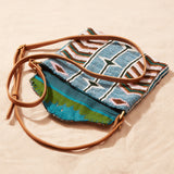 Beaded handbag with geometric pattern and brown leather strap, open showing colorful kanga fabric lining on a sand background