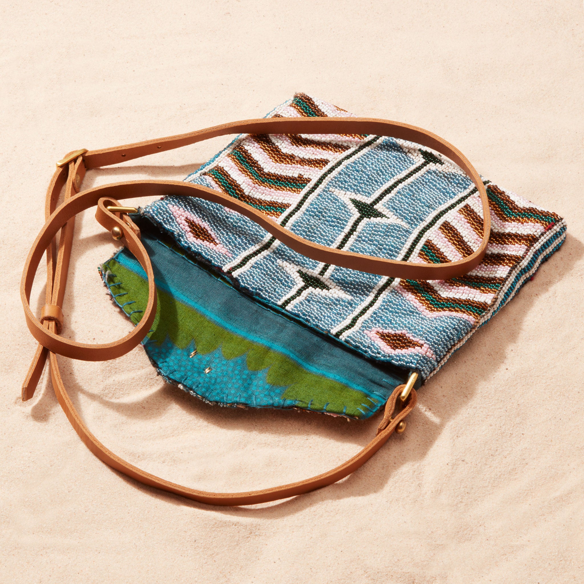 Beaded handbag with geometric pattern and brown leather strap, open showing colorful kanga fabric lining on a sand background