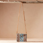 Beaded crossbody bag with a brown leather strap hanging on a branch against a beige background