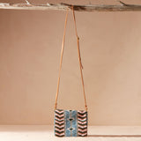 Beaded crossbody bag with a brown leather strap hanging on a branch against a beige background