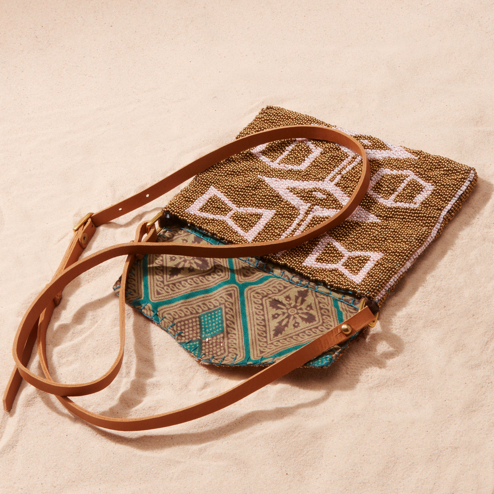 Beaded pink and gold handbag with geometric patterned design and brown leather strap open showing colorful kanga fabric lining on a sand surface