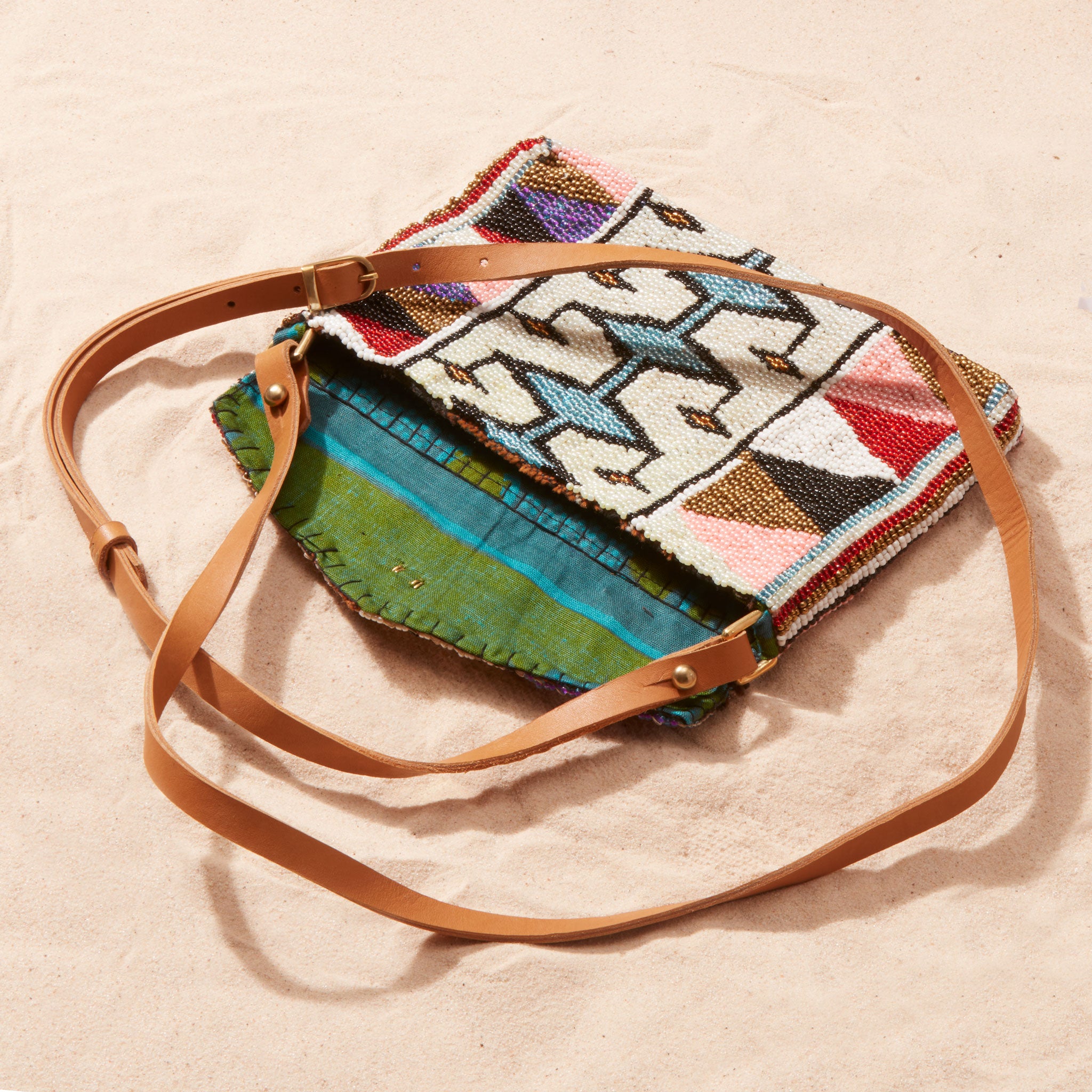 Colorful beaded handbag with a leather strap open with a colorful kanga fabric lining on a sand background