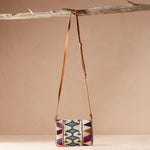 Colorful beaded bag with geometric pattern with a brown leather strap hanging on a branch with a beige background