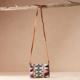 Colorful beaded bag with geometric pattern with a brown leather strap hanging on a branch with a beige background