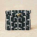 A small beaded clutch bag with a iridescent dark grey and white geometric pattern and a small gold elephant charm on the front, photographed against a beige background.