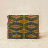 Back view of small beaded clutch bag with a geometric pattern in iridescent bronze and blue with a yellow and pink geometric pattern, featuring a small gold elephant charm on the front flap, set against a neutral beige background.