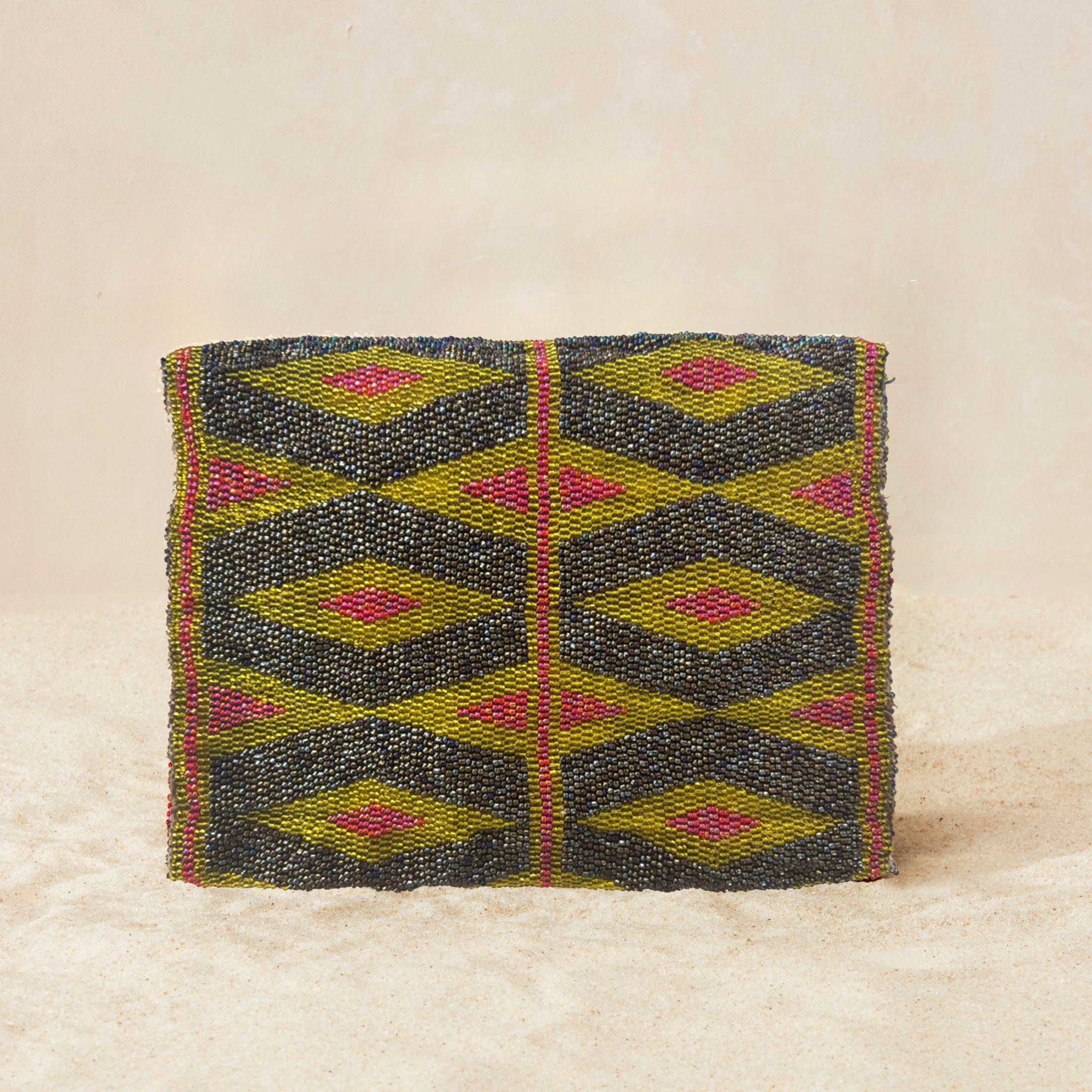 Back view of small beaded clutch bag with a geometric pattern in iridescent bronze and blue with a yellow and pink geometric pattern, featuring a small gold elephant charm on the front flap, set against a neutral beige background.