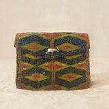 A small beaded clutch bag with a geometric pattern in iridescent bronze and blue with a yellow and pink geometric pattern, featuring a small gold elephant charm on the front flap, set against a neutral beige background.