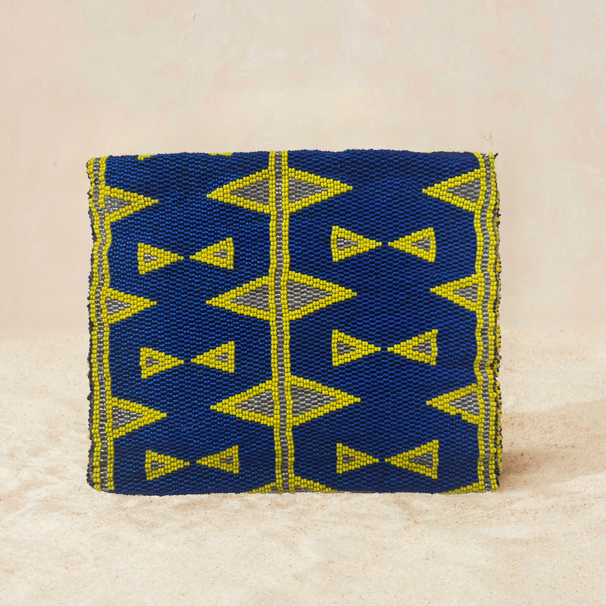Back view of small beaded clutch bag with a blue and yellow geometric pattern, featuring a small elephant charm on the front flap, set against a neutral beige background.