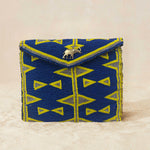 A small beaded clutch bag with a blue and yellow geometric pattern, featuring a small elephant charm on the front flap, set against a neutral beige background.