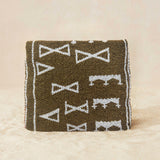 Back view of a small beaded clutch bag with a bronze and white geometric pattern and a small gold elephant charm on the front flap, photographed on a light beige sand surface.