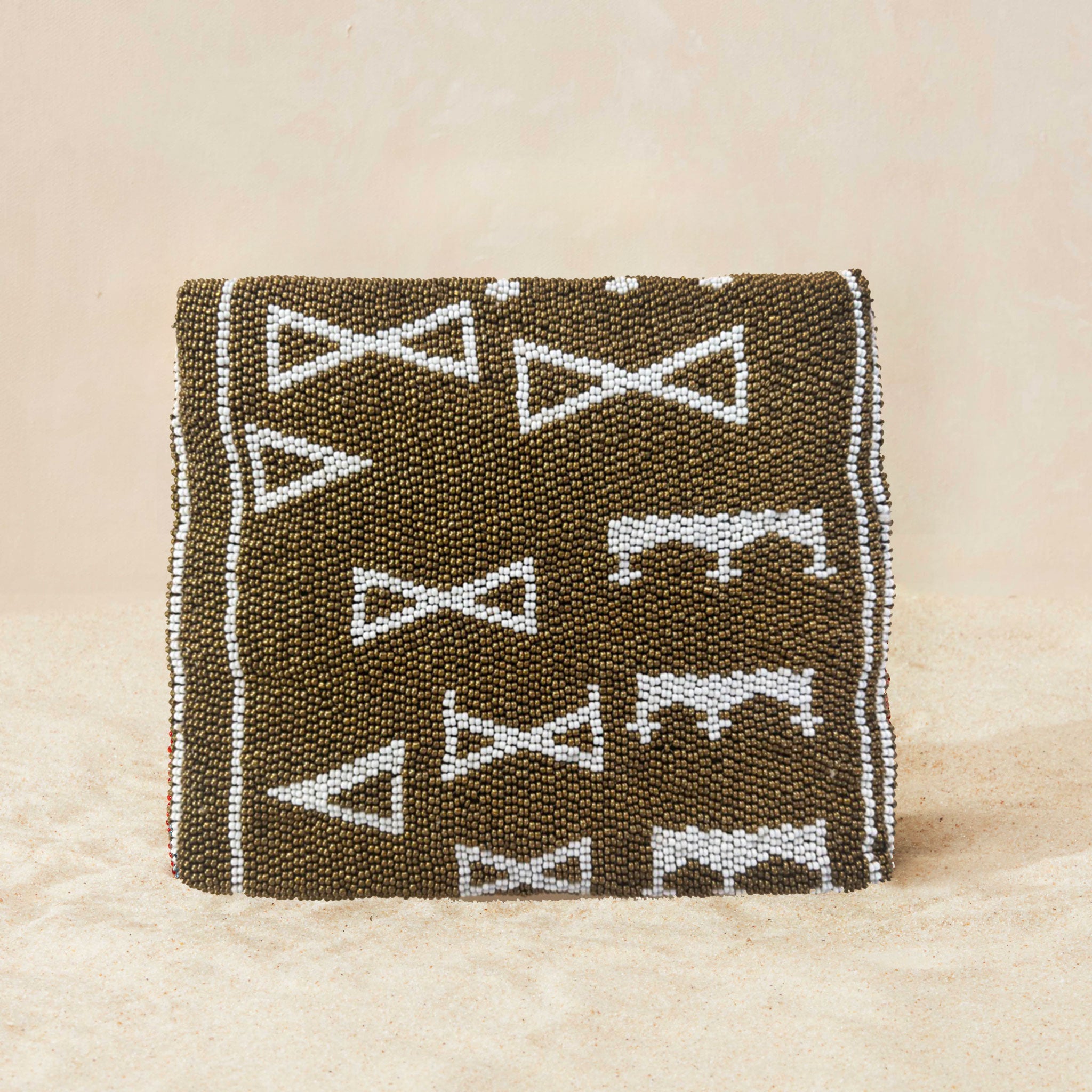 Back view of a small beaded clutch bag with a bronze and white geometric pattern and a small gold elephant charm on the front flap, photographed on a light beige sand surface.