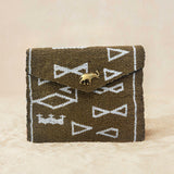 A small beaded clutch bag with a bronze and white geometric pattern and a small gold elephant charm on the front flap, photographed on a light beige sand surface.