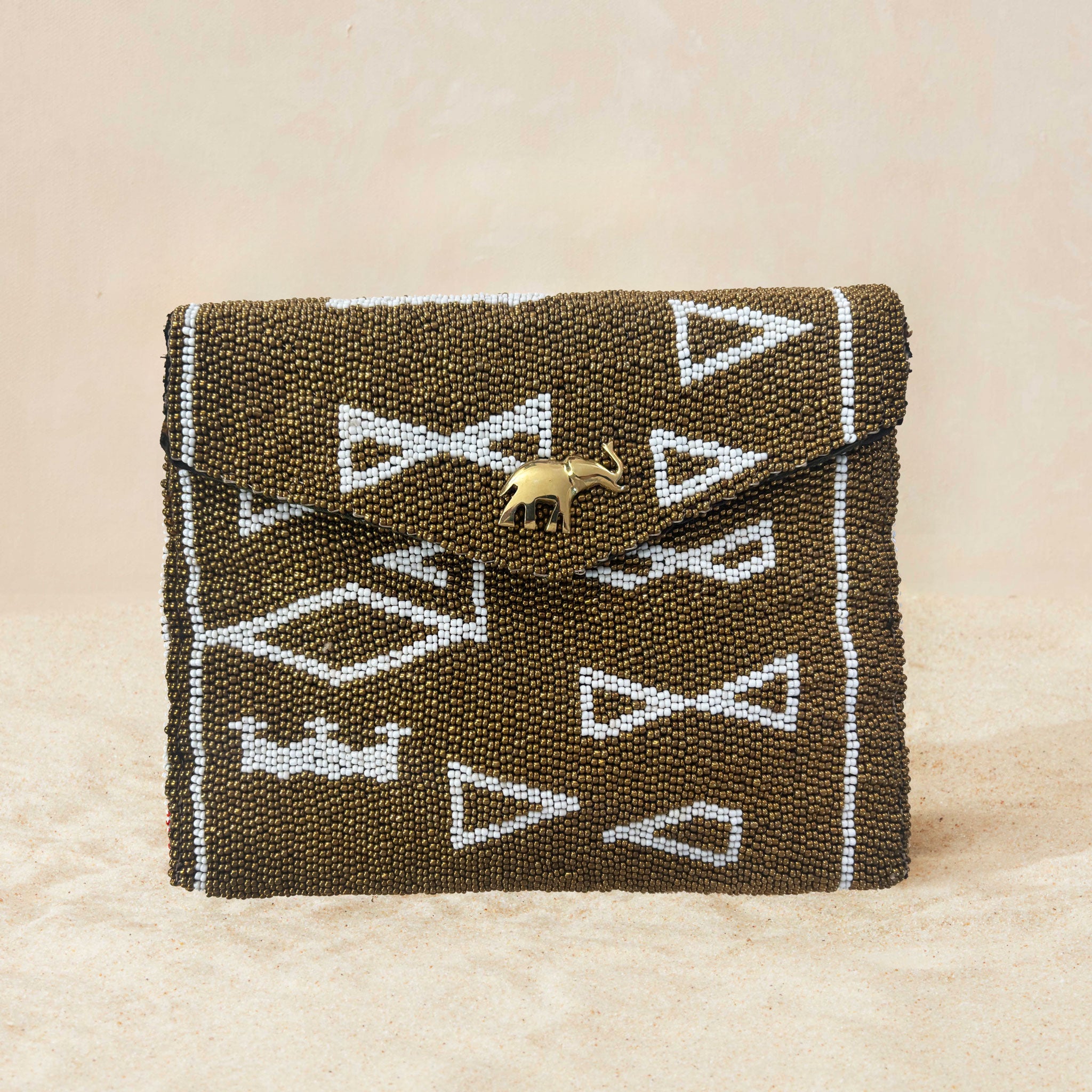 A small beaded clutch bag with a bronze and white geometric pattern and a small gold elephant charm on the front flap, photographed on a light beige sand surface.