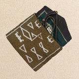 Interior view of a small beaded clutch bag with a bronze and white geometric pattern, featuring a green, mint, and brown kanga fabric lining and a brown leather crossbody strap, photographed on a light beige sand surface.